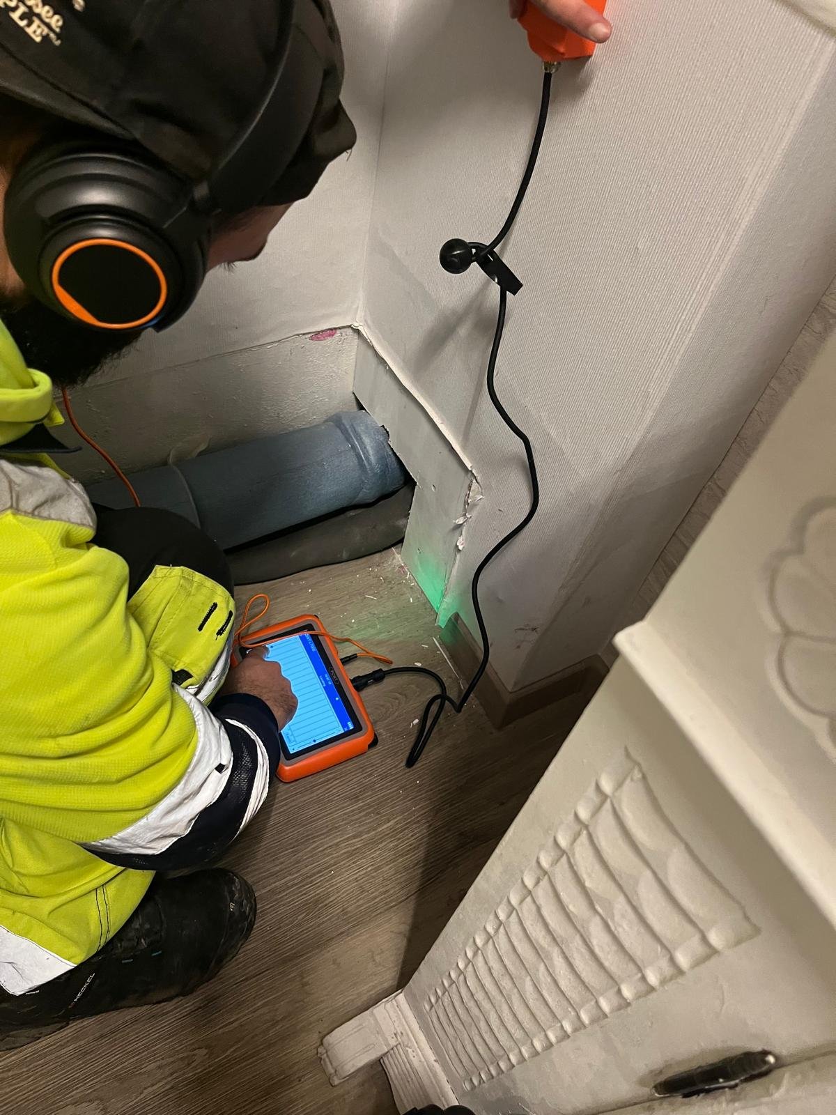 Techniker kniet vor einer geöffneten Wand und misst mit Horchsonde und Tablet ein Rohr zur akustischen Leckortung.