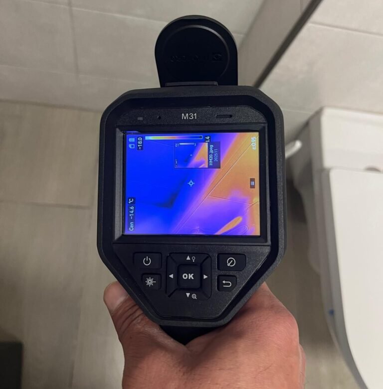 Hand hält eine M31-Wärmebildkamera, die Temperaturunterschiede an einer Badezimmerwand zur Leckortung anzeigt.